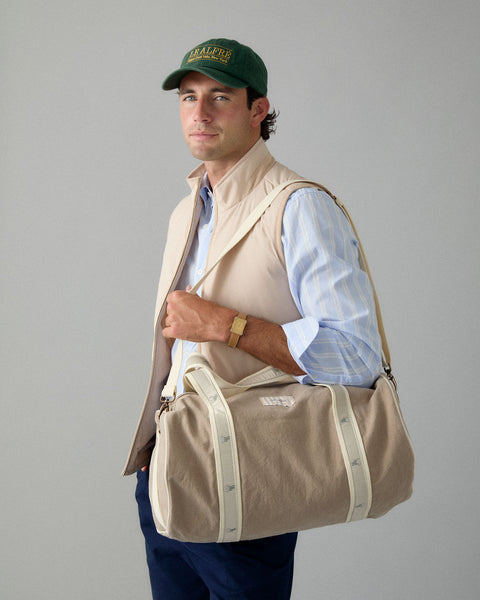Man wearing a beige vest and holding a beige duffel bag against a gray background