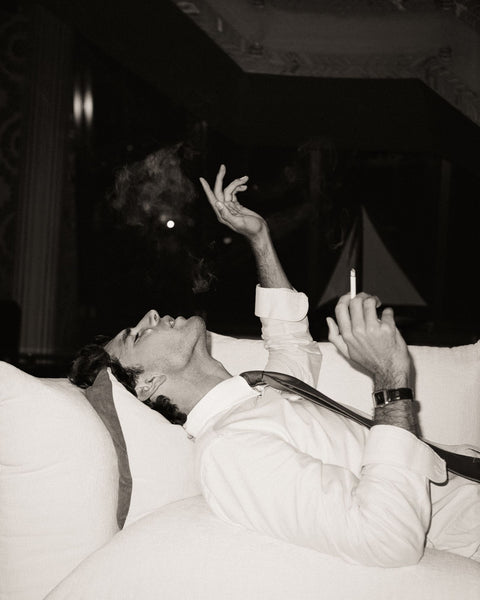 Person lying on a couch with a cigarette in hand, wearing a white outfit.