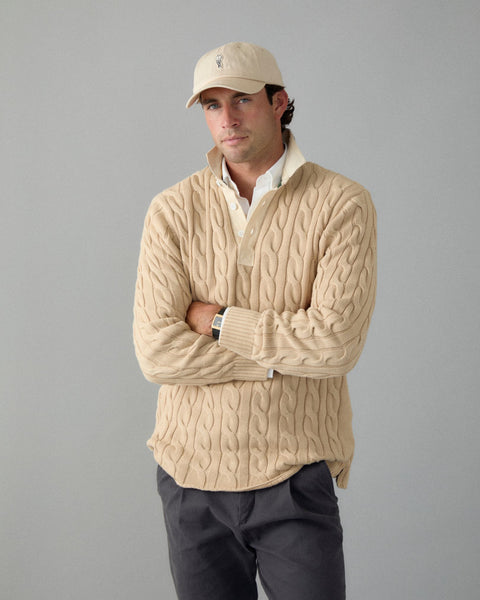 Man wearing a beige cable knit sweater and cap against a gray background