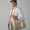 Man wearing a beige vest and holding a beige duffel bag against a gray background