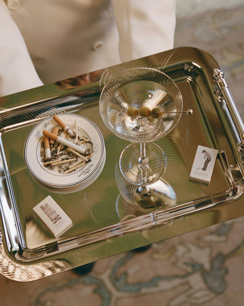 Vintage-style silver tray with martini glass, olives, ashtray with cigarettes, and matchbox, evoking a classic luxury atmosphere.
