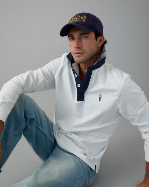 Man wearing a white long-sleeve shirt with navy collar, blue jeans, and a navy cap on a gray background
