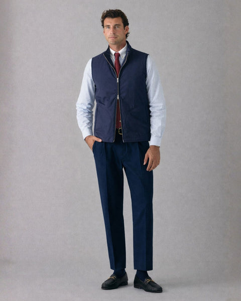 Man wearing a navy vest over a white shirt with a red tie, paired with dark blue pants and shoes on a gray background.