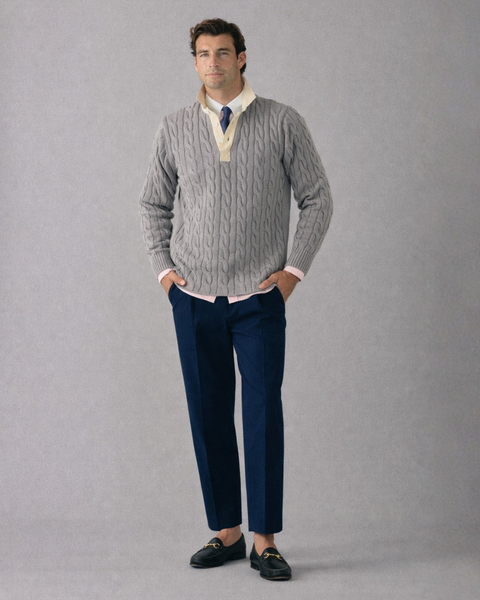 Man wearing a gray cable knit sweater, white shirt, blue pants, and black shoes on a gray background