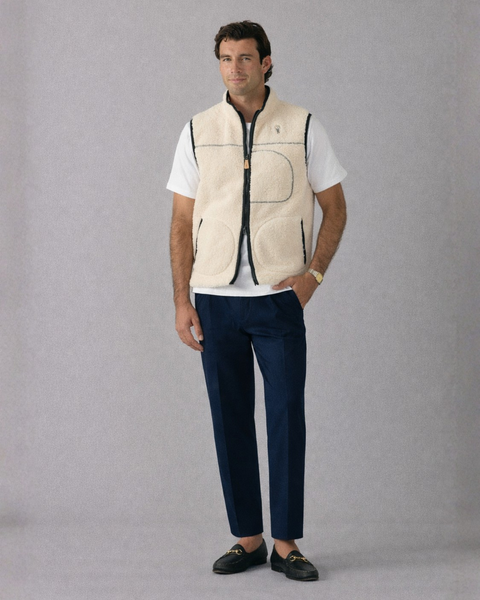 Man wearing a beige vest over a white shirt with navy pants on a gray background
