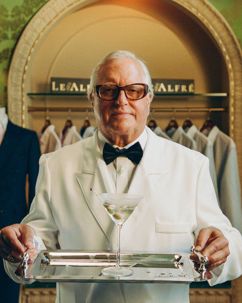 Man in a white suit holding a tray with a cocktail glass in a formal setting.